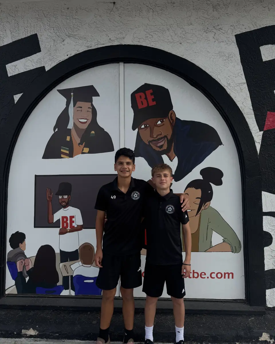 Two students stand arm in arm in front of a Movement BE mural showing a graduate, a teacher at a whiteboard and students in a classroom.