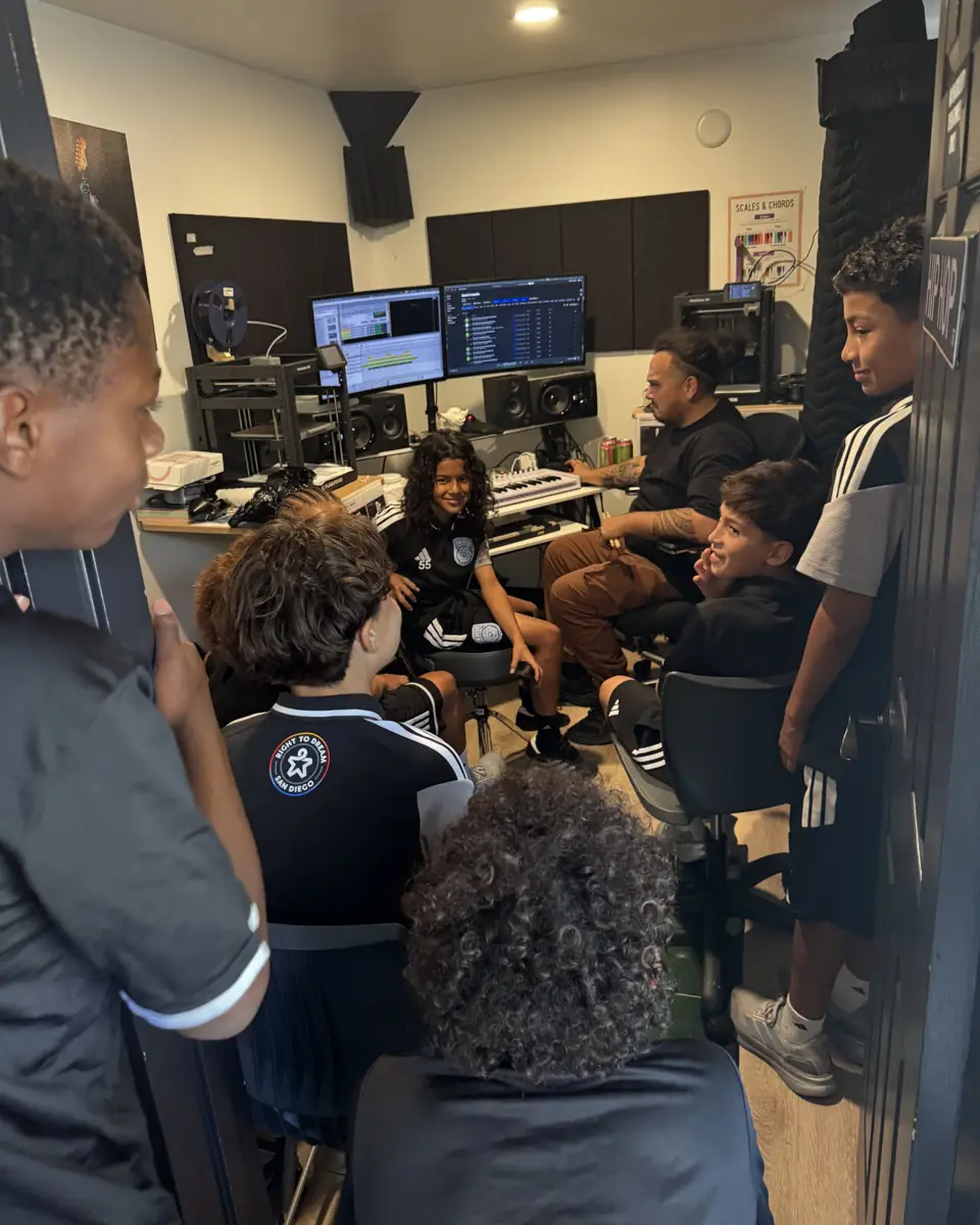 Students crowd into a music production studio as a producer works at the mixing desk and a student sits at the keyboard.