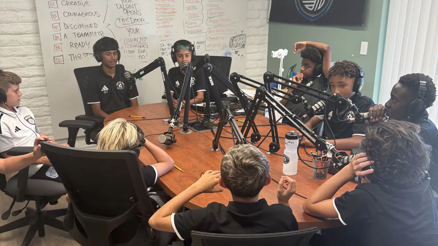 Students in headphones gather around a studio table of podcast microphones, in front of a whiteboard of storytelling notes.
